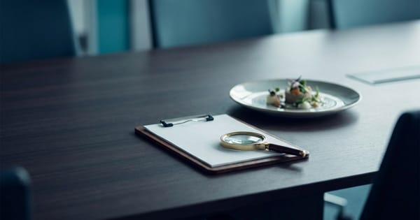 A plain clipboard and a magnifying glass beside an elegantly plated dish in soft focus